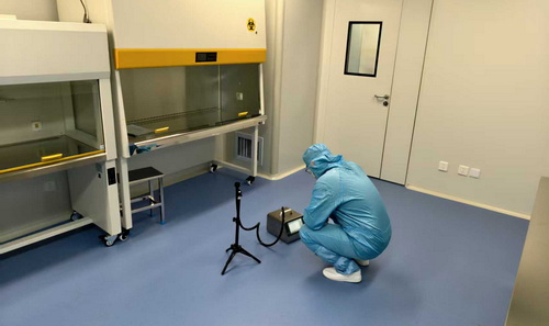 洁净室检测现场，第三方检测机构工程师在生物安全柜旁使用便携式粒子计数器进行悬浮粒子采样，蓝色洁净服，蓝色 PVC 地板，白色围护结构。